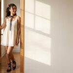 Woman in a cream lace-trim coquette dress with a black ribbon choker and ballet flats standing in sunlit doorway
